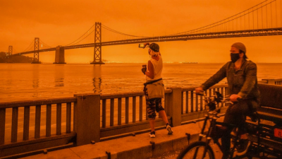 orange photo due to pollution, 2 people standing by a river near a huge bridge, wearing masks.