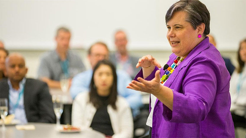 Speaker in a purple jacket presenting to a seated audience in a professional setting.