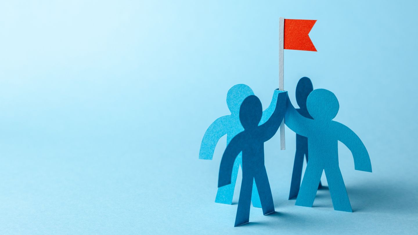 image of 4 paper humans forming a team and holding a flag together