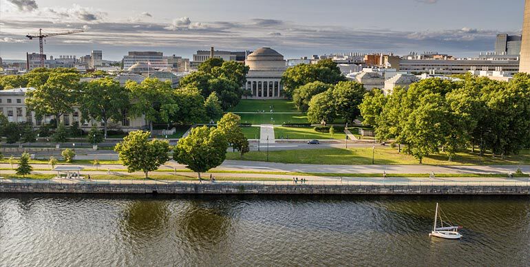 View of MIT dome from across the Charles River with a sailboat in the foreground and green trees lining the riverbank.