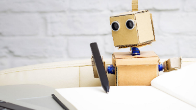 Cardboard robot holding a pen and writing in a blank notebook