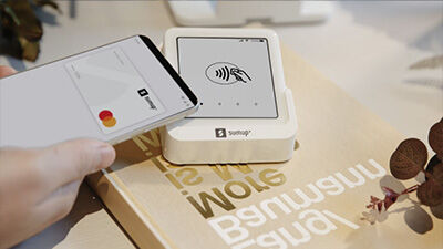 Smartphone making a contactless payment using a white SumUp card reader on a table.