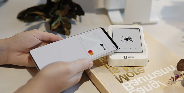 Smartphone making a contactless payment using a white SumUp card reader on a table.
