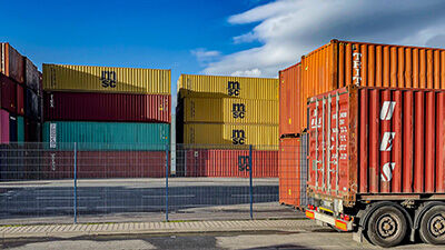 Stacked multicolored shipping containers in an industrial yard with a red truck trailer in the foreground.