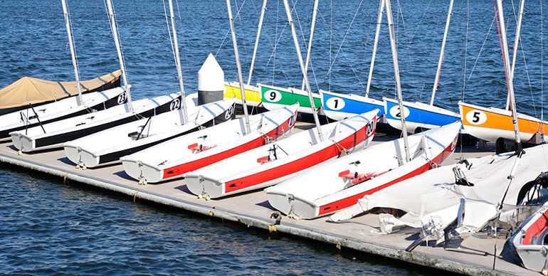 Docked sailboats with numbered sails on the Charles River with the Boston skyline in the background.