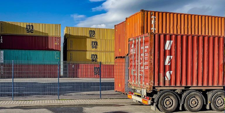 Stacked multicolored shipping containers in an industrial yard with a red truck trailer in the foreground.