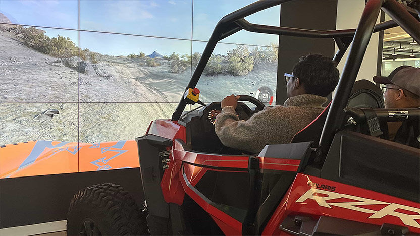 Person using an off-road vehicle simulator in front of a panoramic screen displaying a rugged terrain.