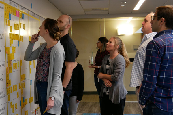 group of colleagues around a white board with post-it notes discussing the flow of work and practicing dynamic work design