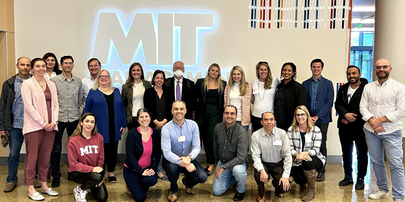 class photo of Communication and Persuasion in the Digital Age participants and faculty - roughly 25 people gathered in the lobby of a campus building in front of a glowing "MIT" sign